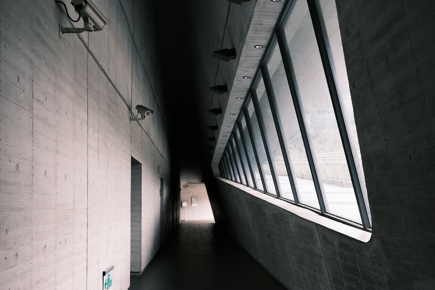 A quiet concrete corridor, empty, lit by a long angled window running the length of one wall.