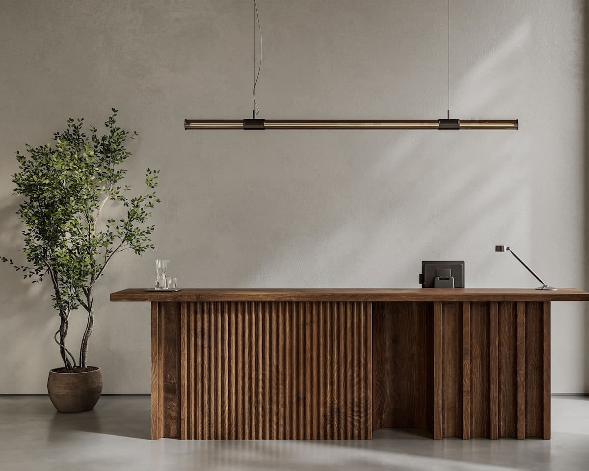A reception desk in fluted walnut against a plaster wall, a linear pendant above it and a potted tree beside it.