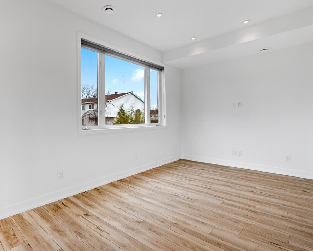 An empty room in a home, pale walls and oak floors, daylight from a window looking onto trees.