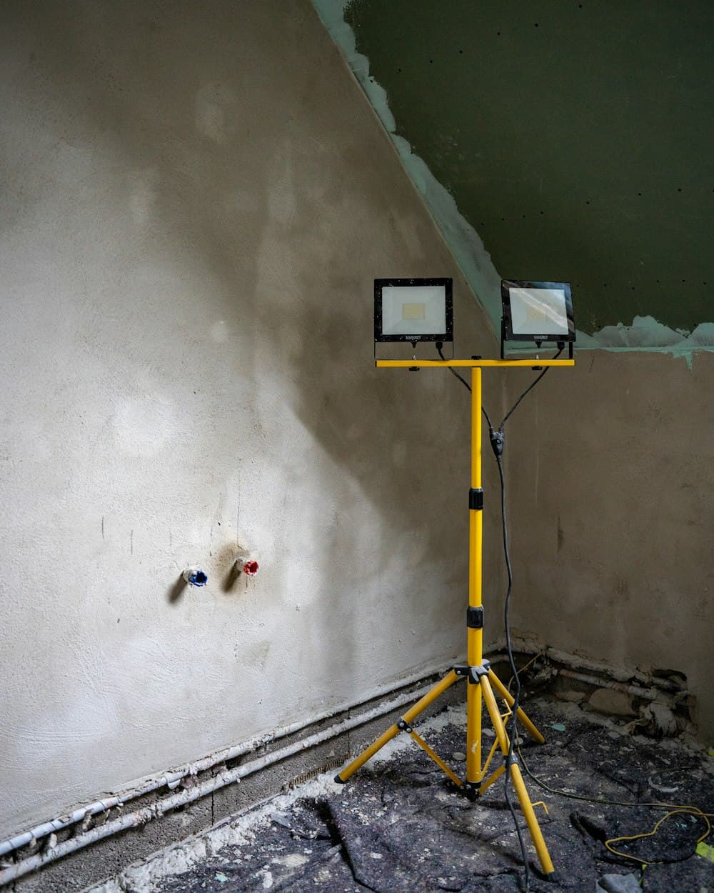 A newly plastered wall with exposed pipe stubs, lit by a yellow work light on a tripod.